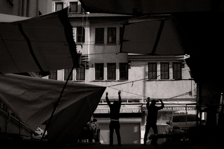 Black and white photo of silhouetted figures adjusting awnings in an urban setting.